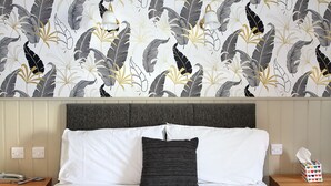 Un lit bien fait, avec du linge de lit blanc et un seul oreiller noir, une table de chevet avec une boîte de mouchoirs, et un mur recouvert d’un papier peint à motifs de feuilles.