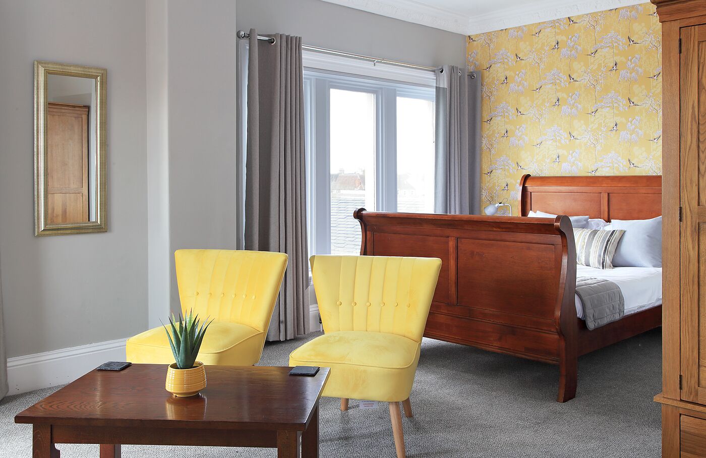 A room with a wooden bed, two yellow armchairs, a wooden table with a plant, and a window with curtains.