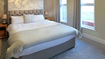A large bed with a tufted headboard, a bedside table with a lamp, and a view of the outdoors through a window.