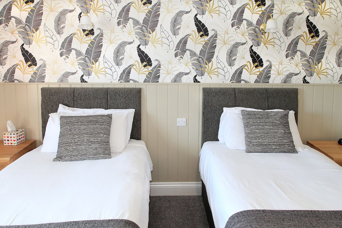 Quarto de hotel com duas camas, cada uma com roupa de cama branca e almofadas cinzentas. O quarto apresenta um papel de parede estampado com motivos de folhas e uma cabeceira de madeira.