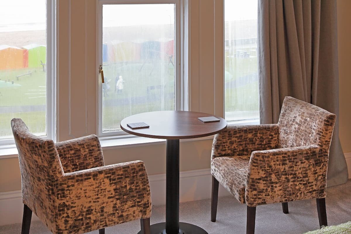 Quarto com duas poltronas estampadas, uma mesa redonda e vista para um campo de esportes através de uma janela.