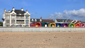 Une rangée de cabanes de plage aux couleurs vives, avec un grand bâtiment historique en arrière-plan.