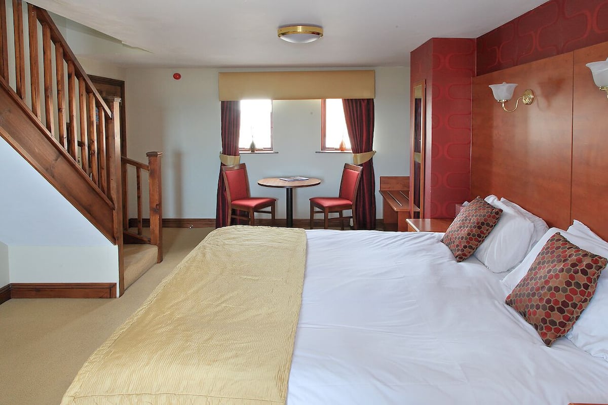 Quarto de hotel com uma cama grande, escada de madeira, pequena mesa de jantar com cadeiras e vista para o exterior por duas janelas.