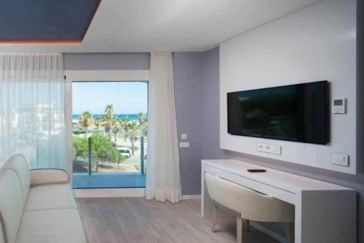A modern hotel room with a bed, a sofa, a TV, and a balcony with a view of the city and palm trees.