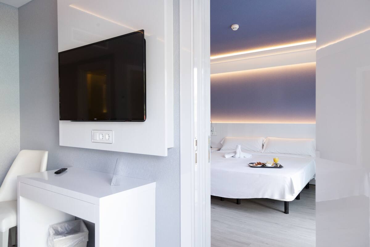 A modern hotel room with a flat-screen TV mounted on the wall, a white desk with a remote control, a bed with white bedding, and a breakfast tray on the bed.