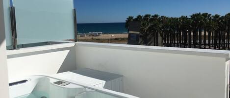A balcony with a hot tub, a view of the beach, and palm trees.
