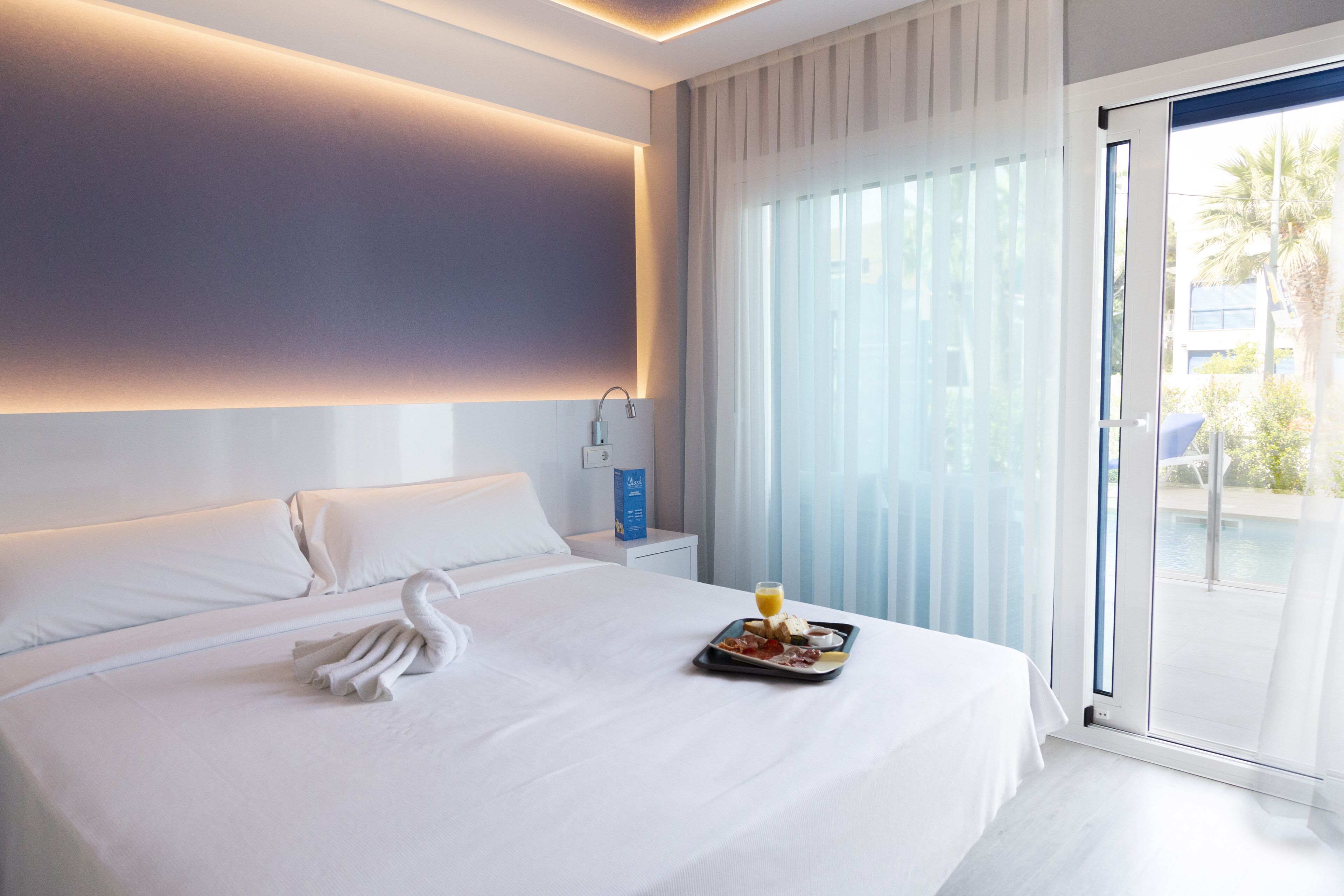 A modern hotel room with a large bed, a tray with breakfast, a swan-shaped towel, and a view of the outdoors through a sliding door.