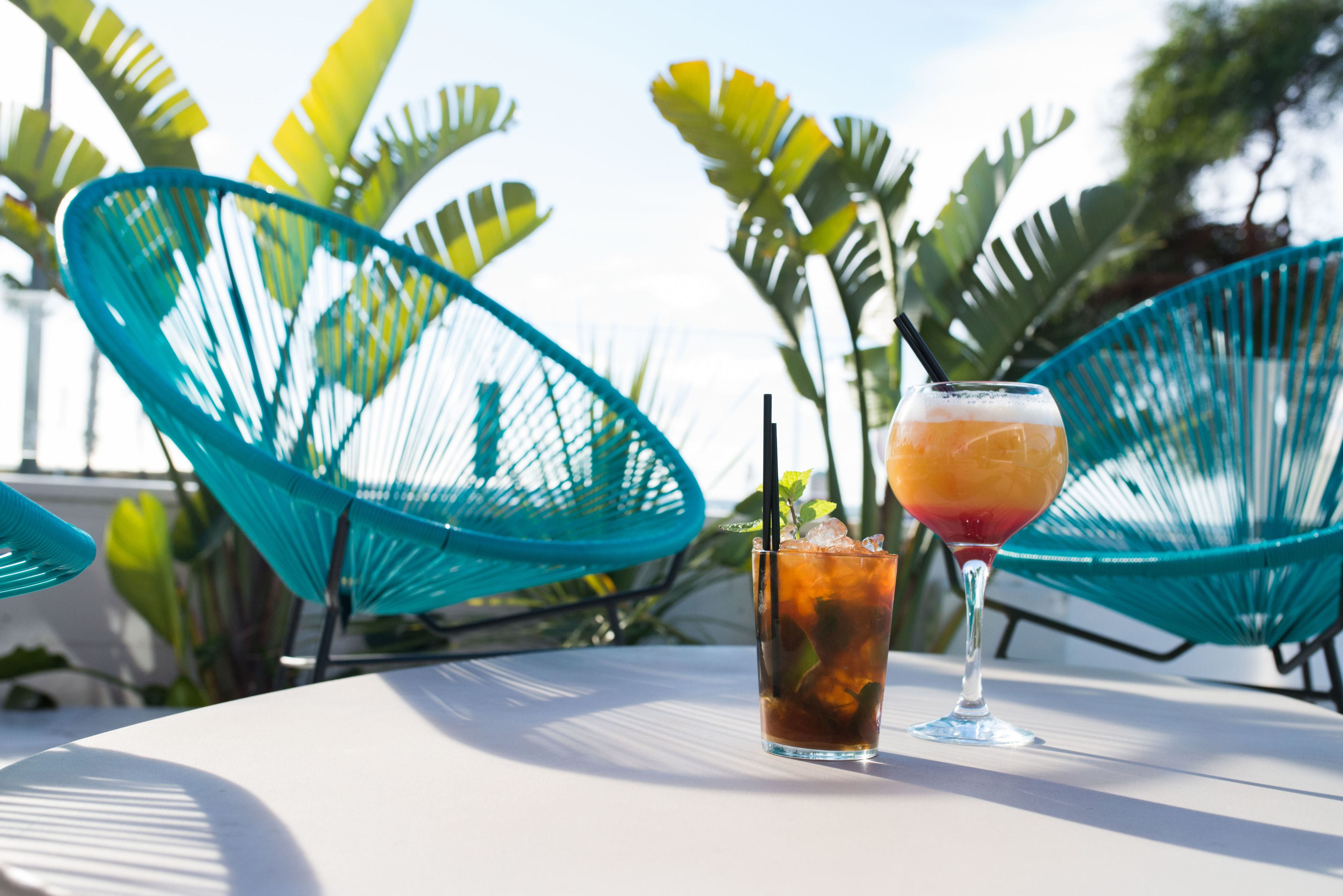 Two drinks on a white table with blue chairs and tropical plants in the background.