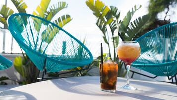 Dos bebidas sobre una mesa blanca con sillas azules y plantas tropicales al fondo.