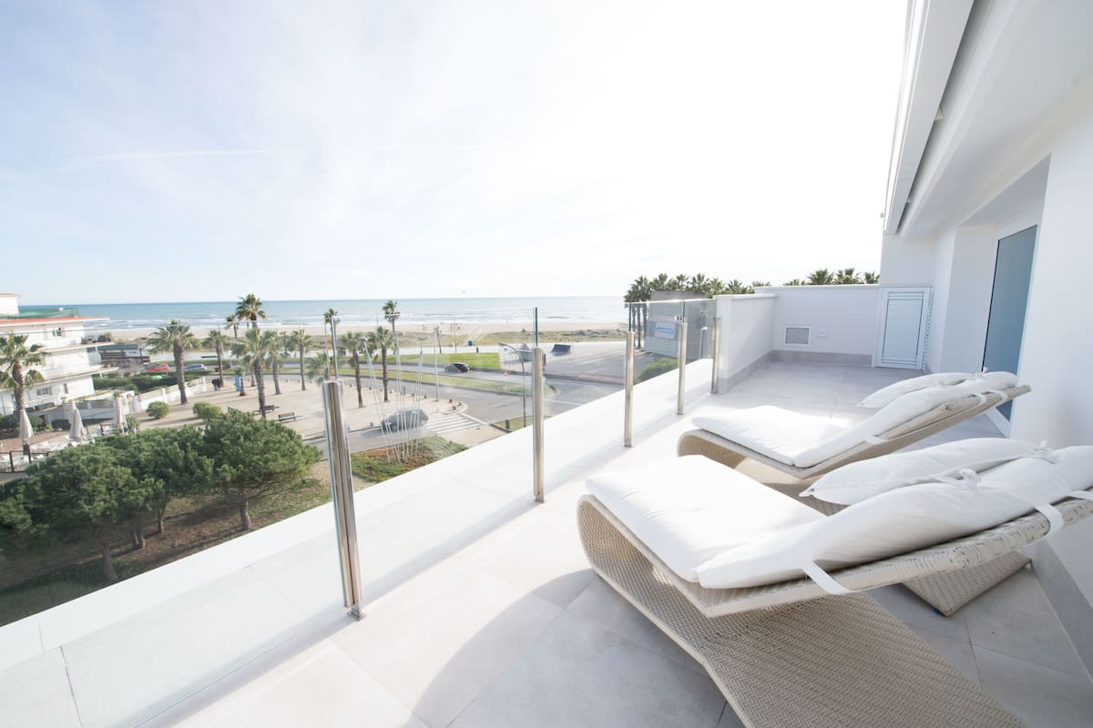 A balcony with a view of the beach, lounge chairs, and a modern building.