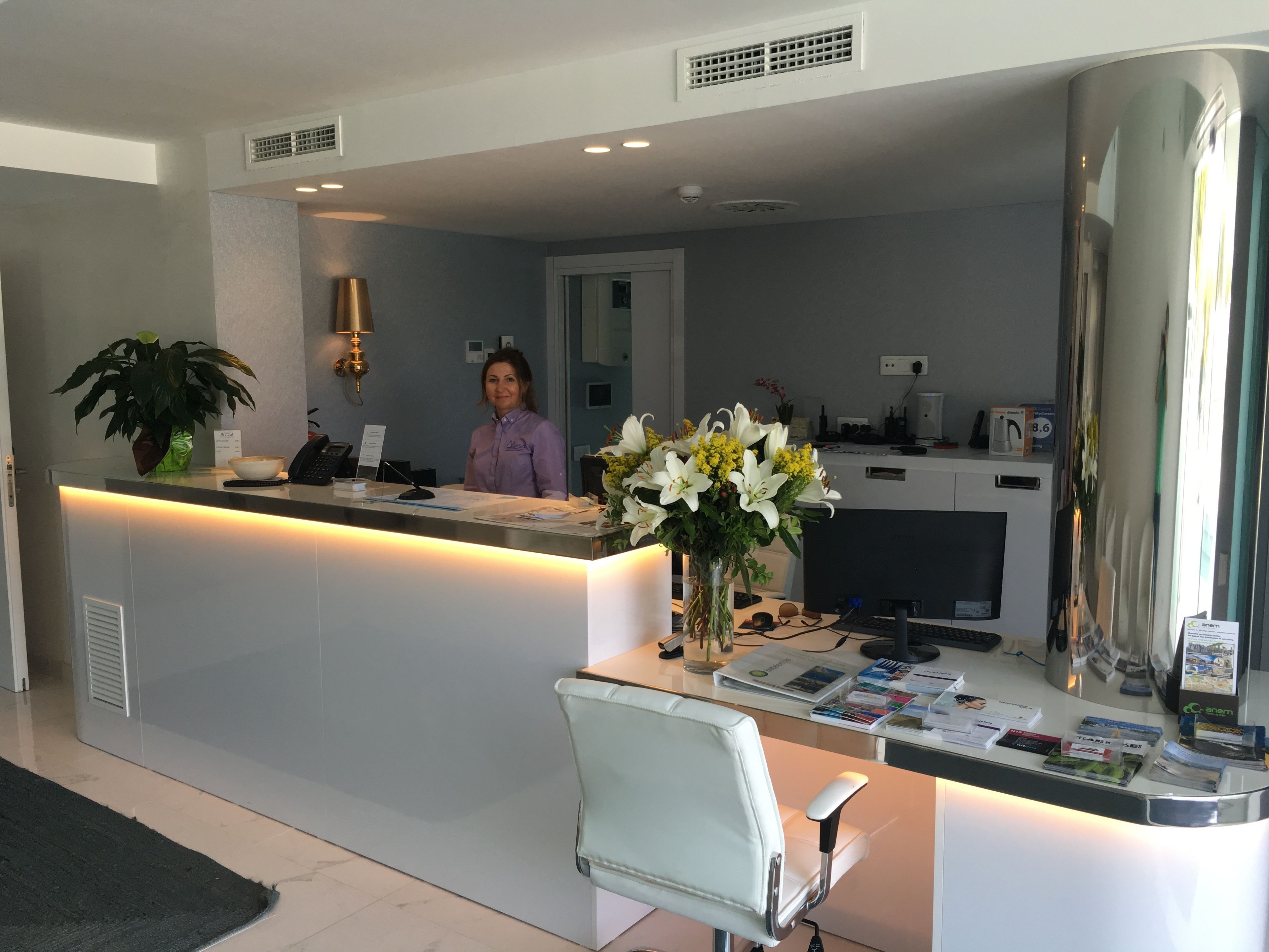 Un mostrador de recepción con una recepcionista, una planta, un jarrón con flores y una computadora.