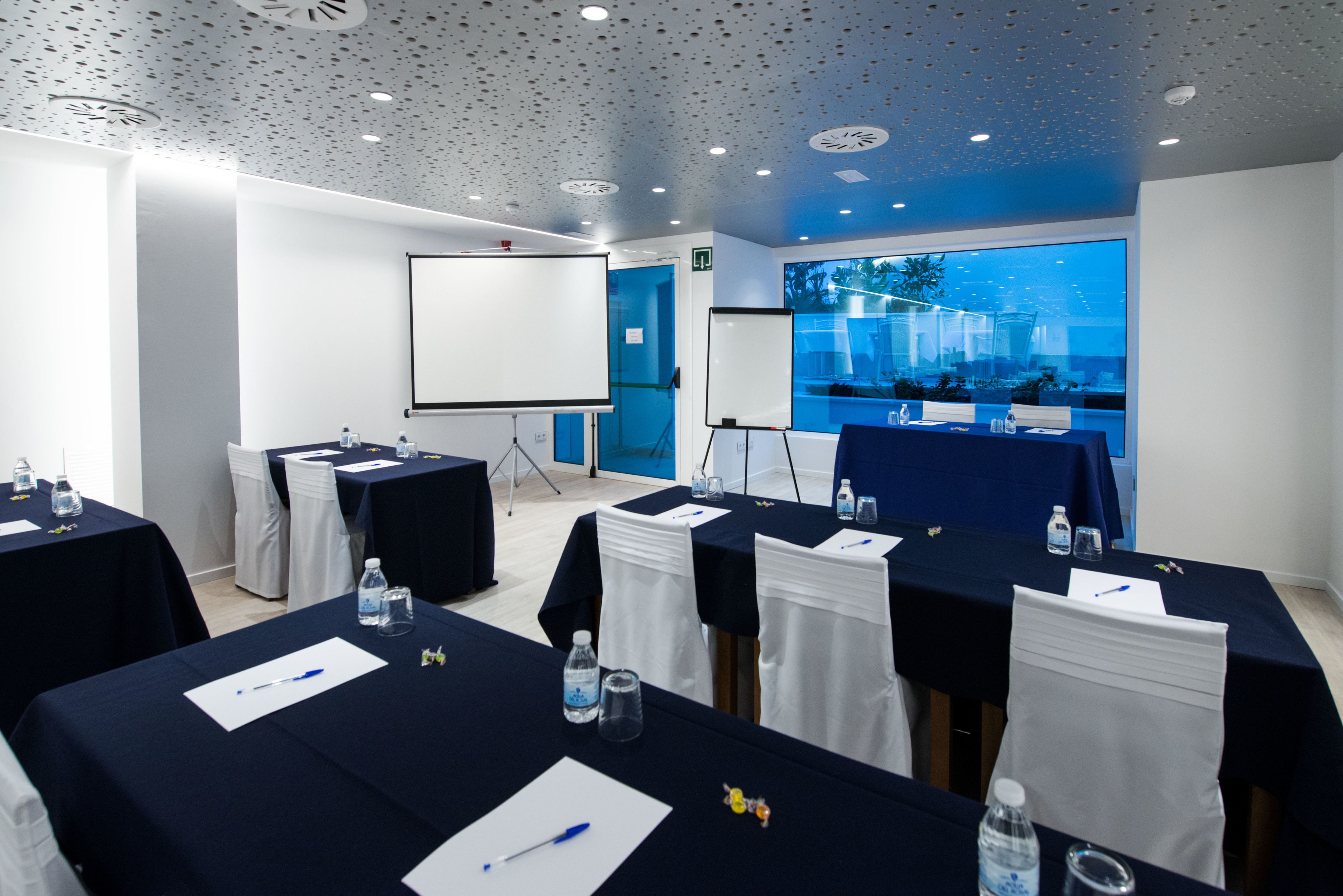 A conference room with tables set for meetings, equipped with water bottles and pens, and a large screen for presentations.