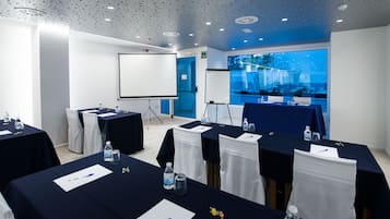 A conference room with tables set for meetings, equipped with water bottles and pens, and a large screen for presentations.