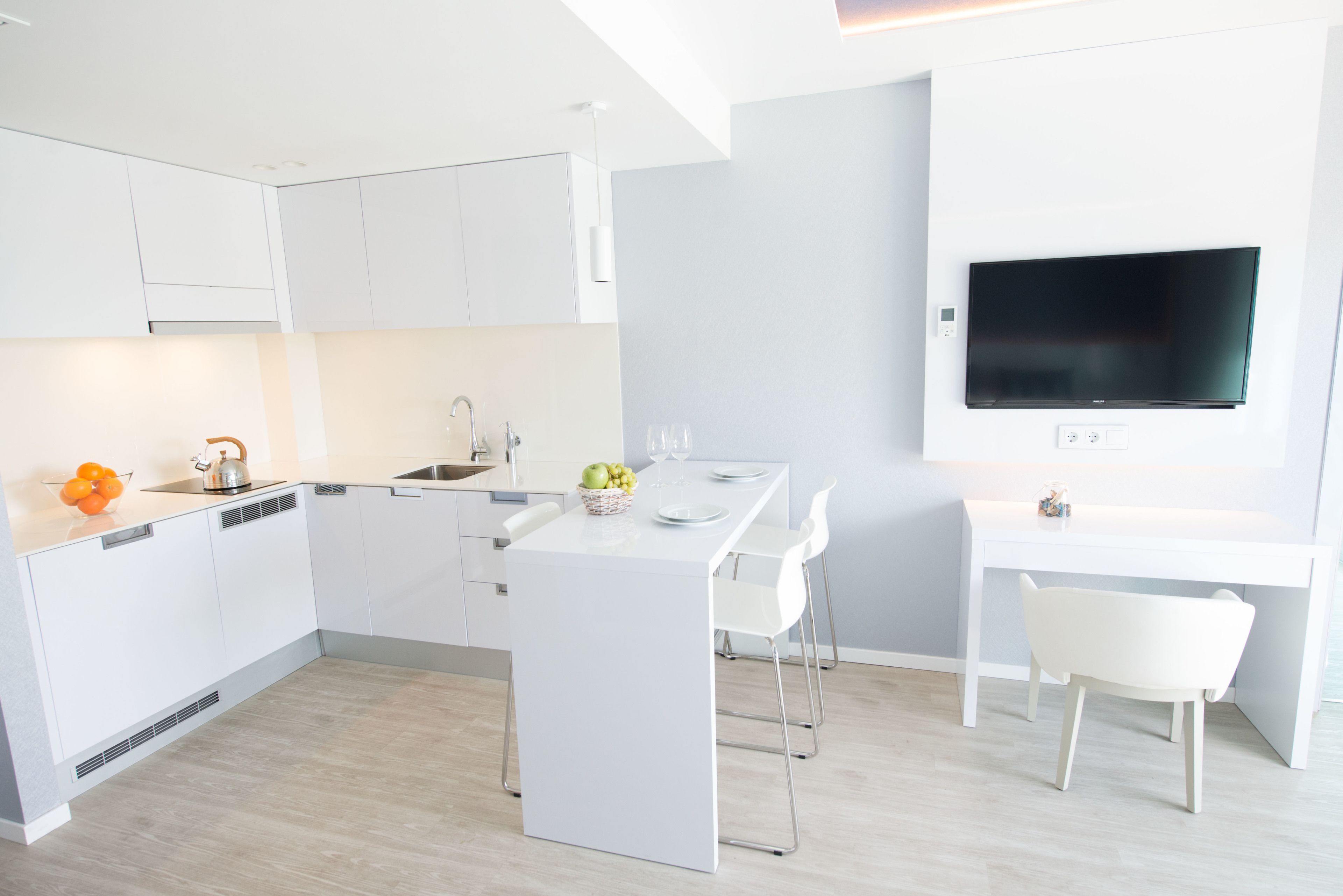 Een moderne keuken met witte kasten, een eethoek met een witte tafel en stoelen, en een tv aan de wand.