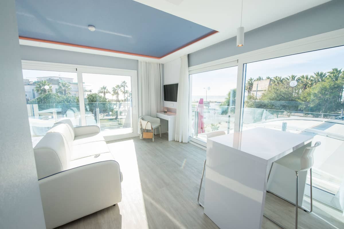 A modern hotel room with a balcony, a white sofa, a dining table, and a TV.