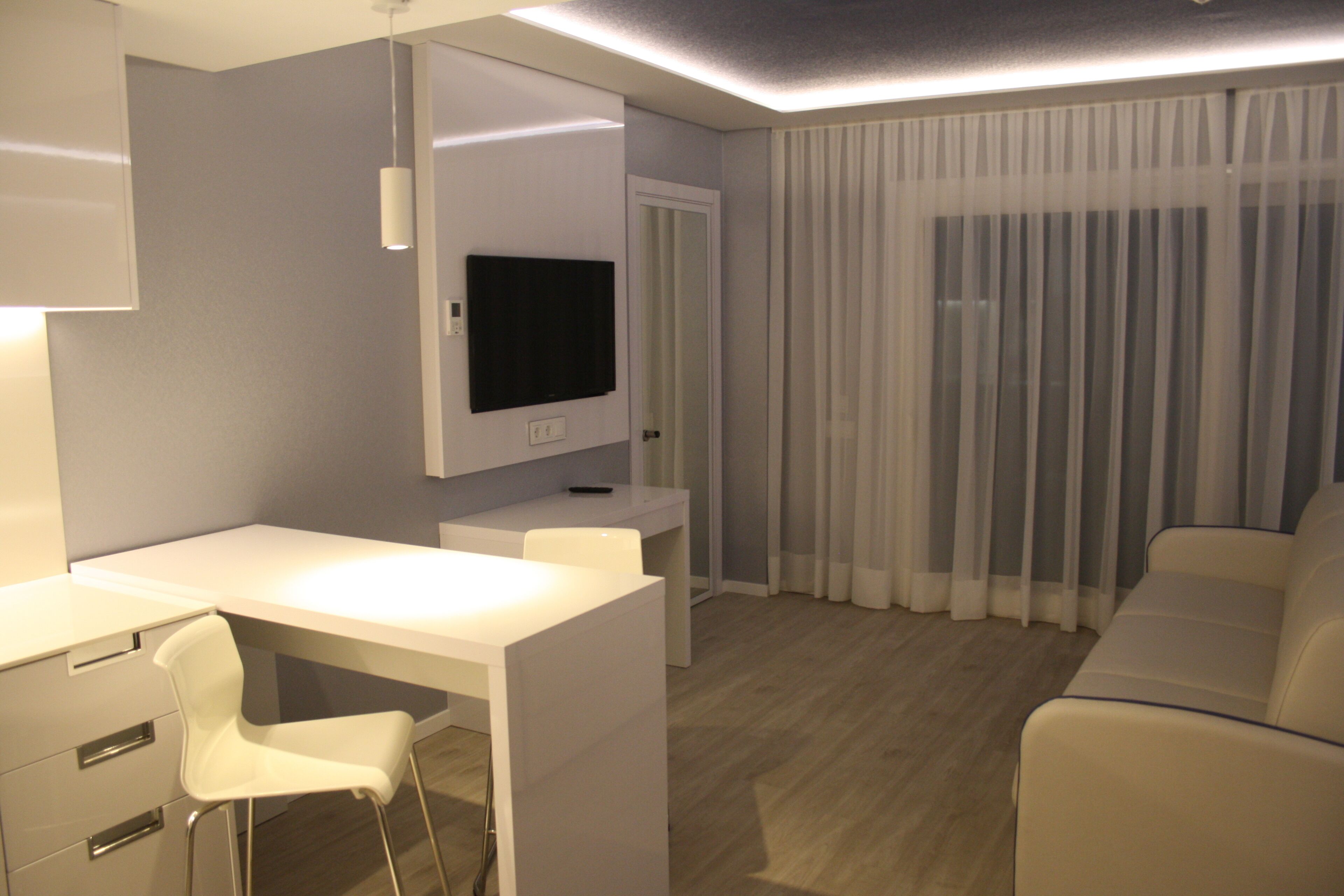 Ein modernes Hotelzimmer mit einem wandmontierten Flachbildfernseher, einem weißen Schreibtisch mit Stuhl, einer Sitzecke und einem großen Fenster mit durchsichtigen Vorhängen.