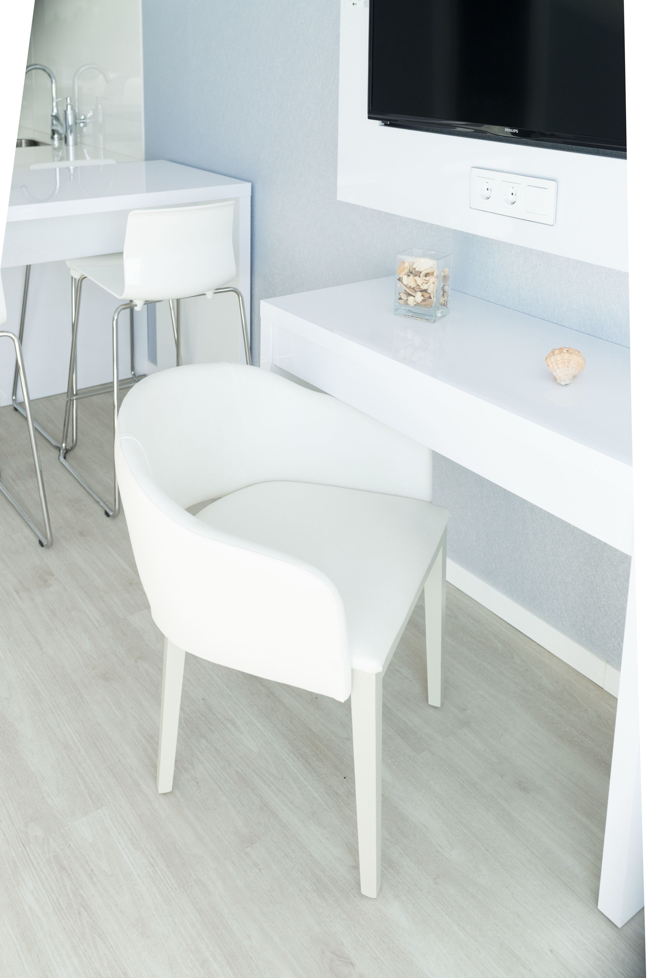 A modern, minimalist room with a white desk, a white chair, a television mounted on the wall, and a small decorative item on the desk.