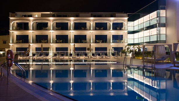 Un hotel de varios pisos con una zona de piscina, que incluye una piscina grande, tumbonas y una fachada bien iluminada.
