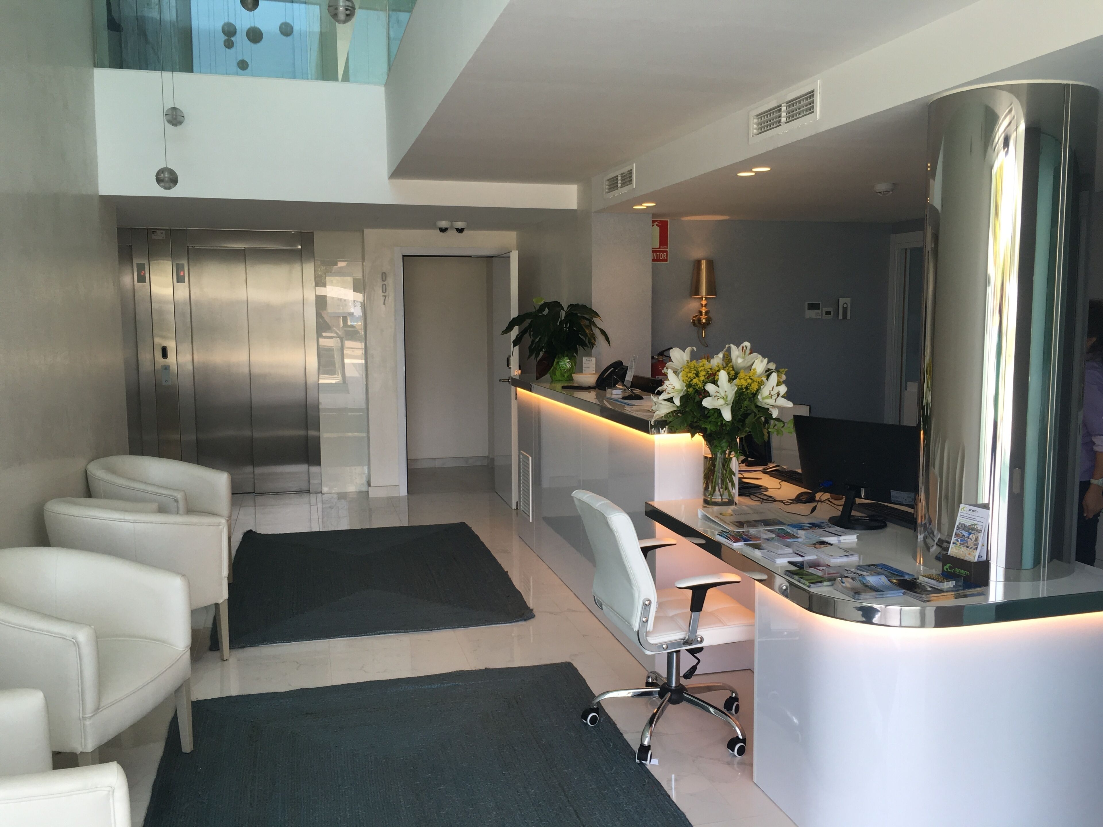 A hotel lobby with a reception desk, a seating area with chairs, an elevator, and a potted plant.