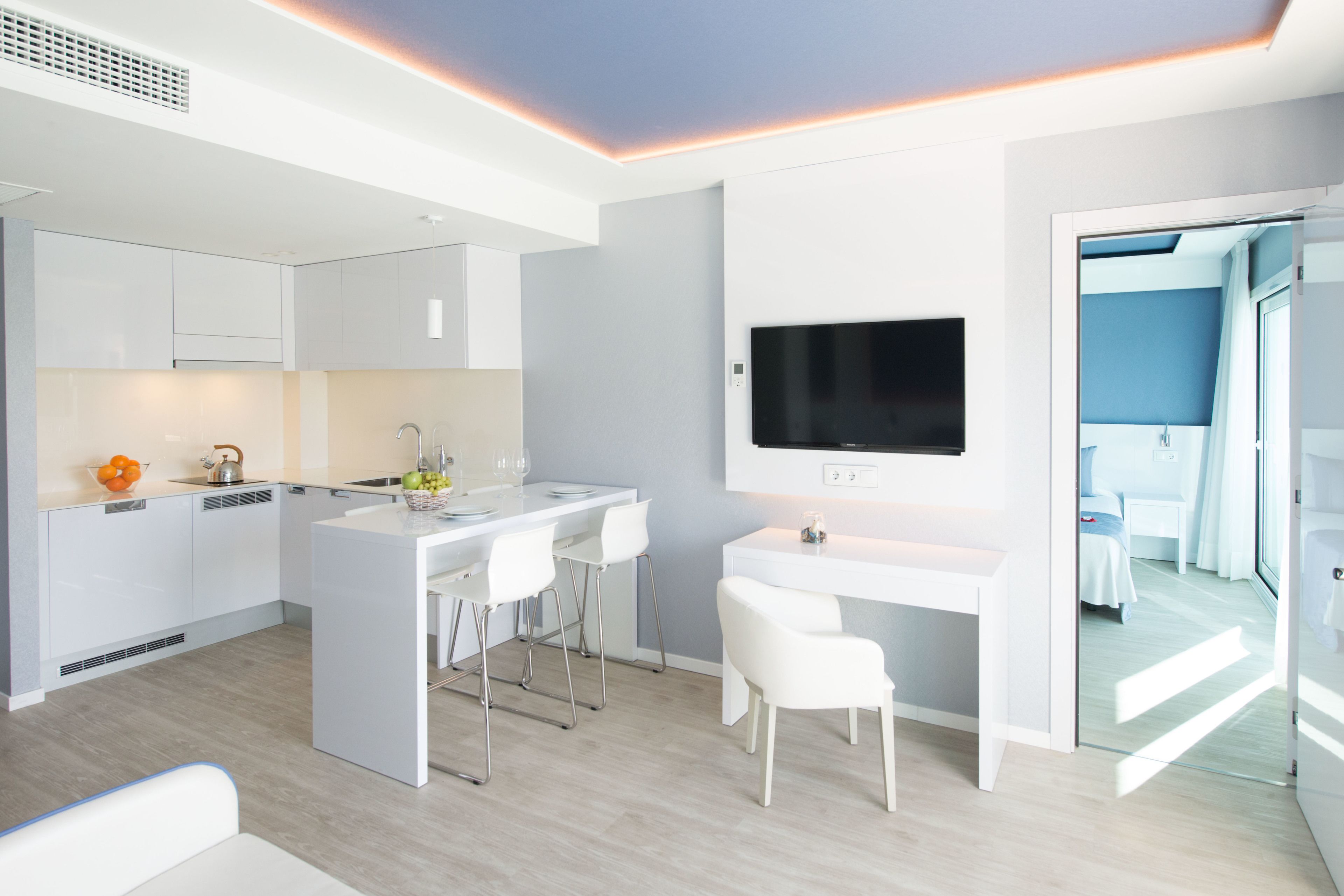 A modern kitchen with white cabinetry, a central island with bar stools, a flat-screen TV mounted on the wall, and a view into a bedroom through an open door.