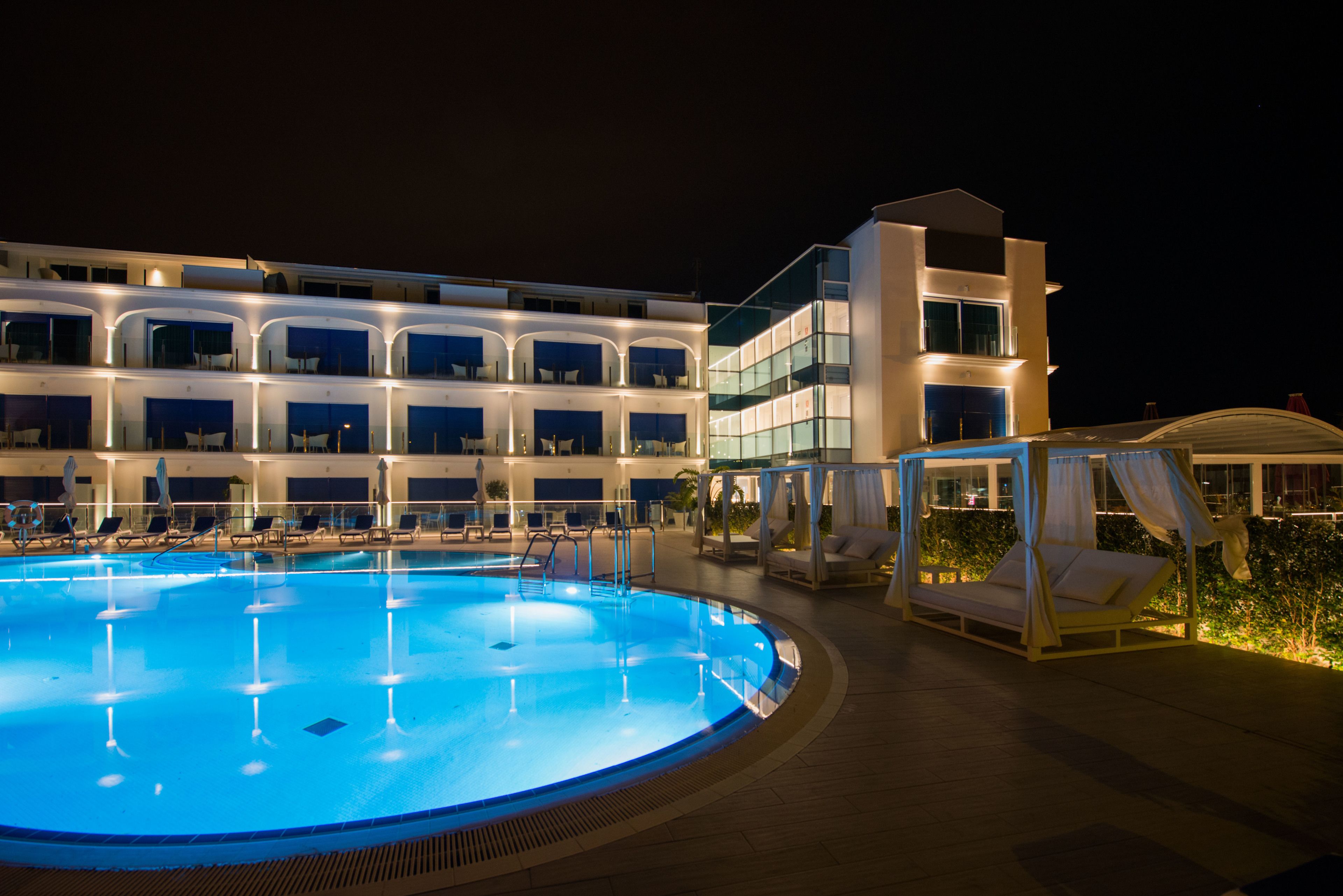 A swimming pool with lounge chairs and a canopy bed, surrounded by a hotel building with multiple balconies and large windows.