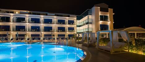 A swimming pool with lounge chairs and a canopy bed, surrounded by a hotel building with multiple balconies and large windows.