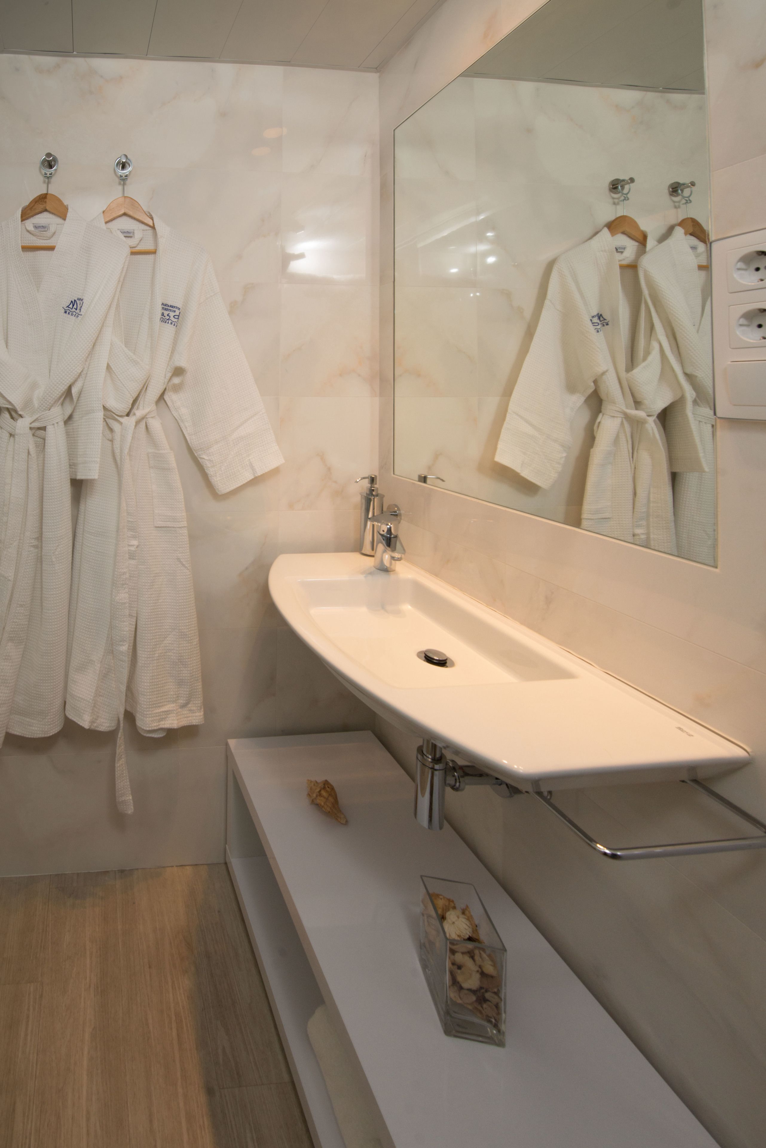 Un baño moderno con tocador de mármol, un espejo grande y dos albornoces blancos colgados en un perchero de madera.