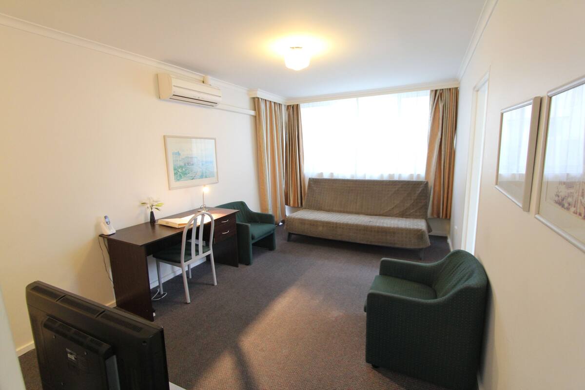 A compact hotel room with a desk, a green armchair, a sofa, and a large window with curtains.