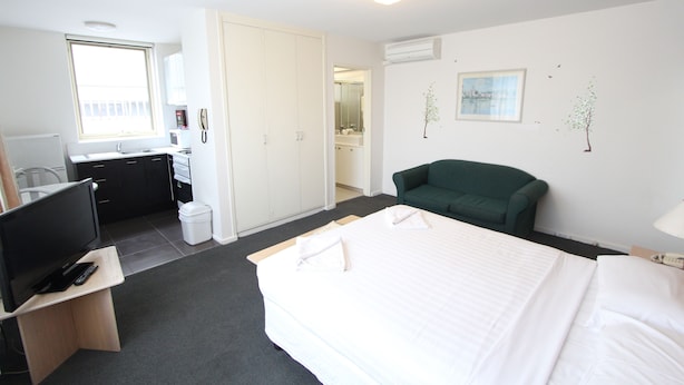 A hotel room with a bed, a desk with a TV, a green chair, and a bathroom visible through an open door. - Drummond Apartments Services (Carlton)
