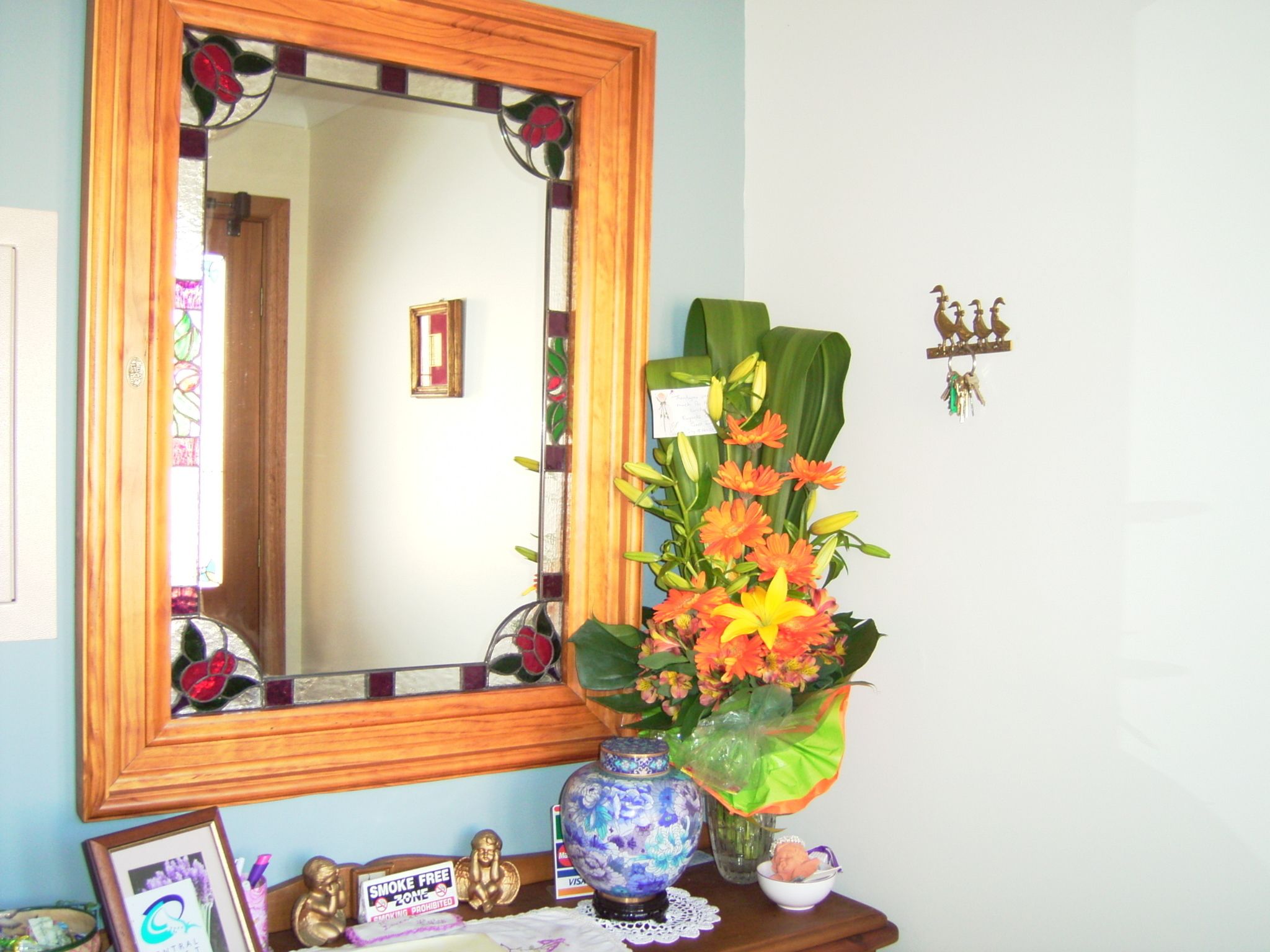 A mirror with a wooden frame, a vase with flowers, a small bowl, and a framed picture on a wooden surface.