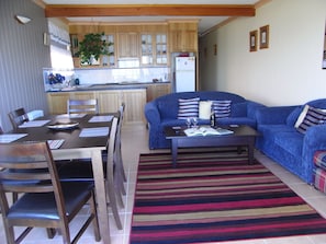 A living room with a blue sofa, a coffee table with a book and a bottle, and a striped rug. There is a dining table with chairs and a kitchen in the background.