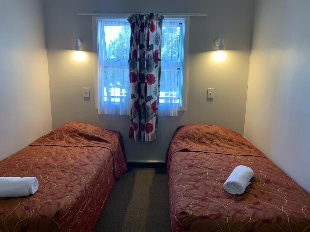 A room with two single beds, each with a red patterned bedspread and a rolled white towel on top. There is a window with a floral curtain and two wall-mounted lights.