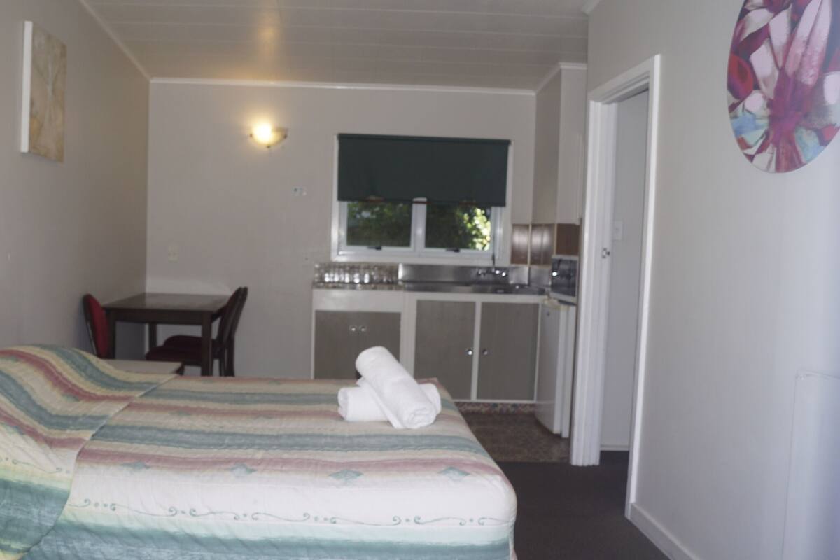A compact room with a bed, a small table, a chair, a kitchenette, and a door.
