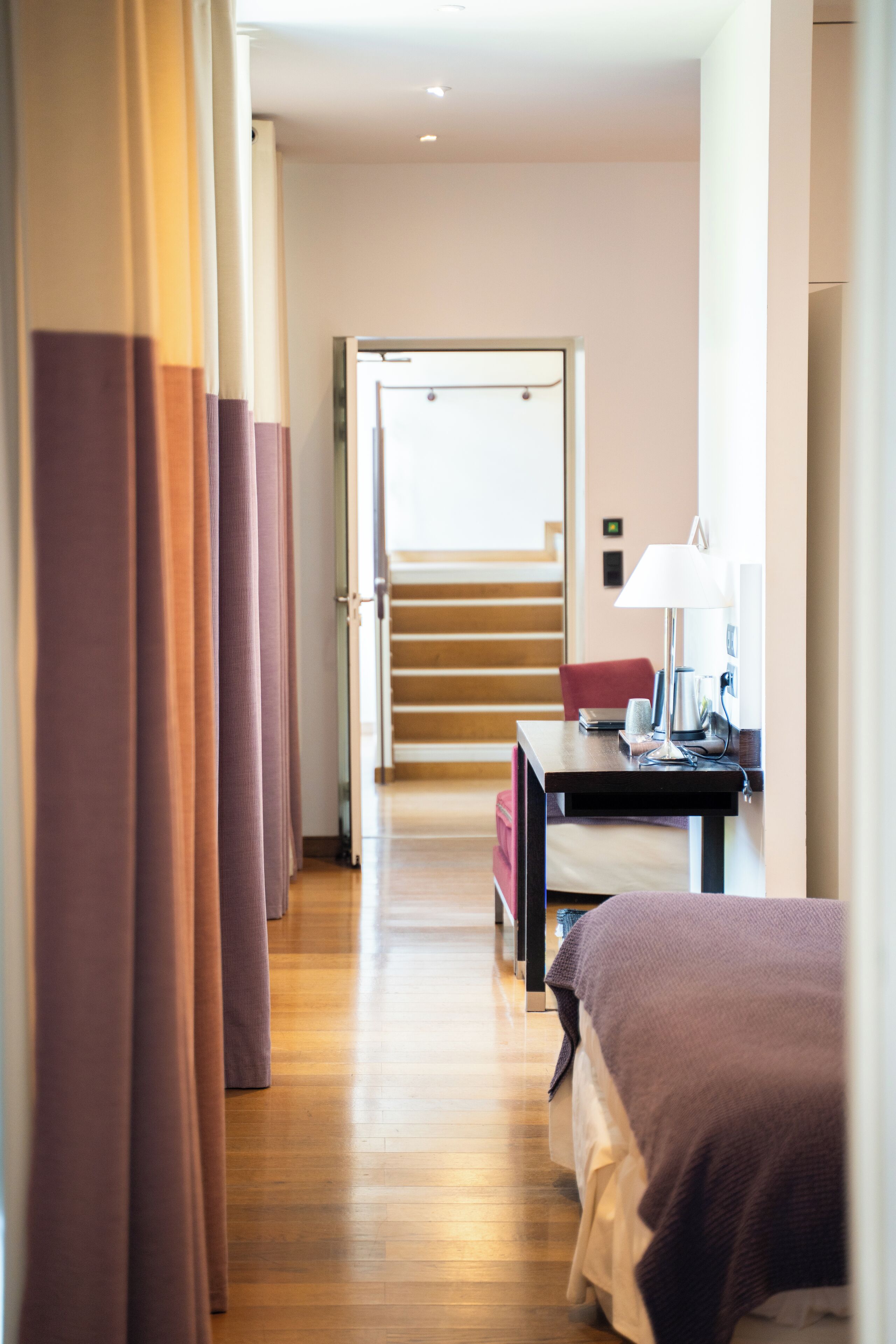 Una habitación de hotel con piso de madera, una cama, un escritorio con lámpara y vista a una escalera que lleva a otra habitación.