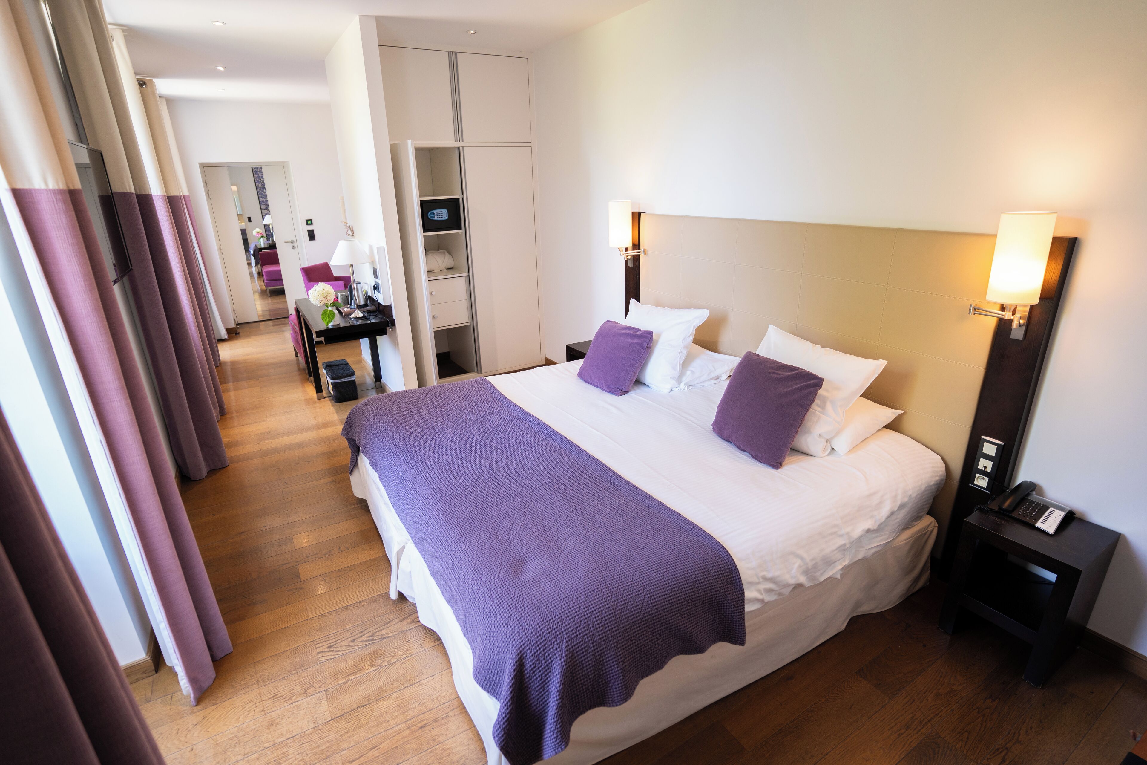A hotel room with a large bed, a bedside table with a lamp, a wardrobe, and a desk with a computer.