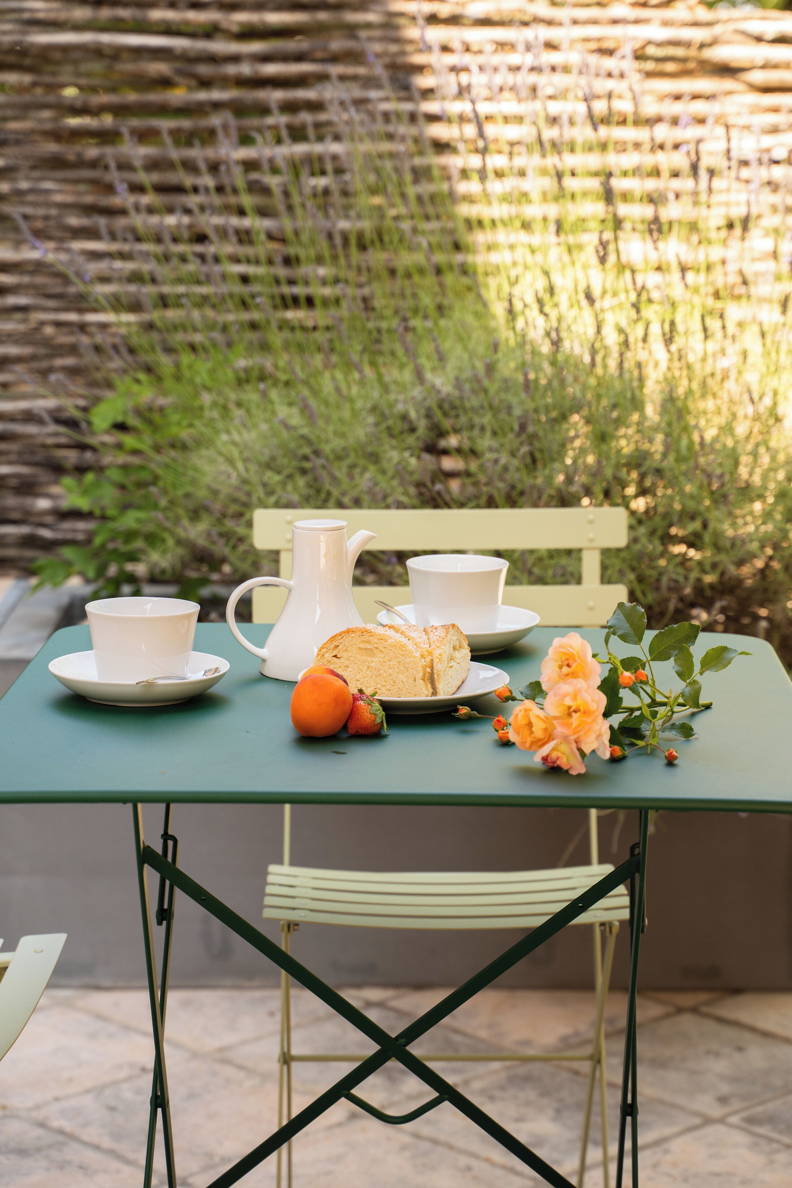 Une table avec une carafe, des tasses, du pain, des fruits et des fleurs, disposée à l’extérieur, avec un banc et de la verdure en arrière-plan.