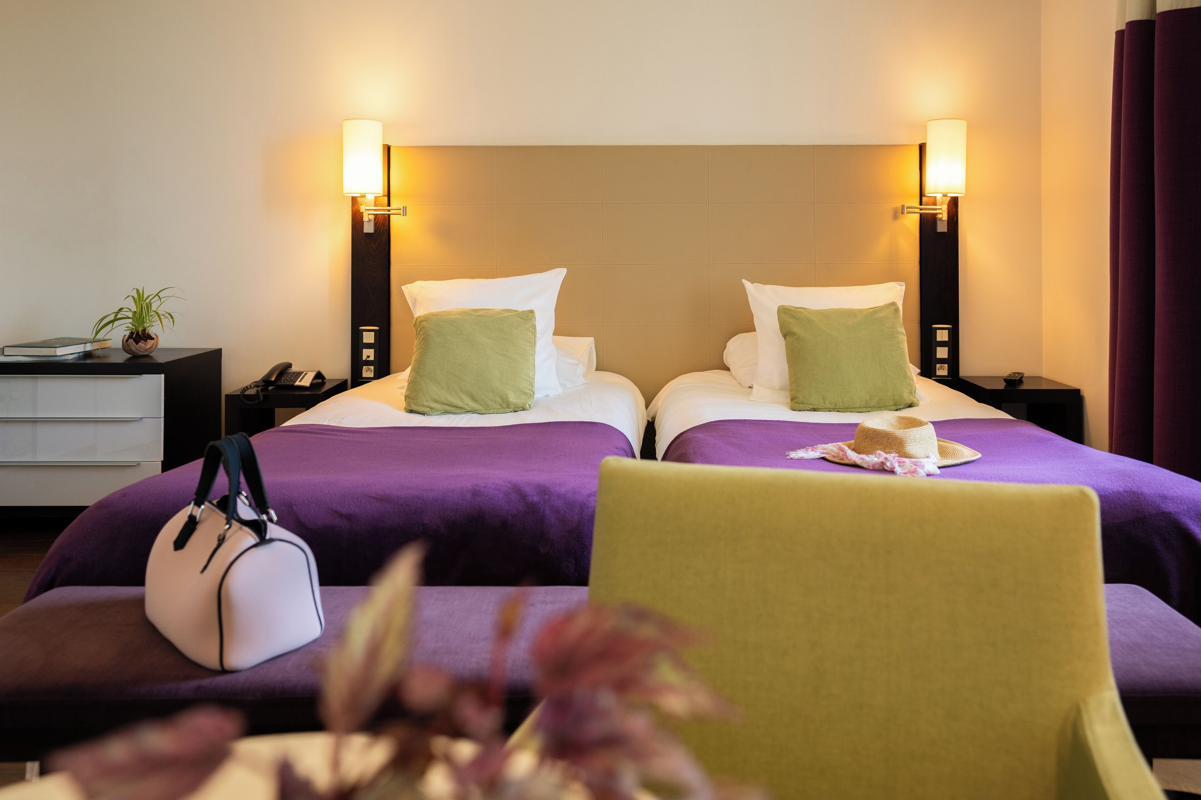 A hotel room with two beds, each with a pillow and a bedside table with a lamp. There is a chair with a hat on it and a handbag on the floor.