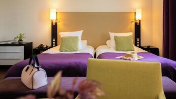 A hotel room with two beds, each with a pillow and a bedside table with a lamp. There is a chair with a hat on it and a handbag on the floor.