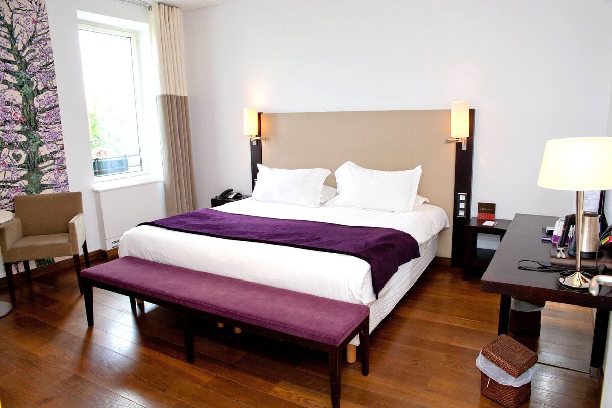 A hotel room with a large bed, a bench, a desk, a chair, and a nightstand.