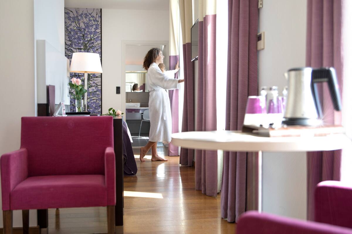 A hotel room with a wooden floor, a pink armchair, a table with a lamp, and a TV.