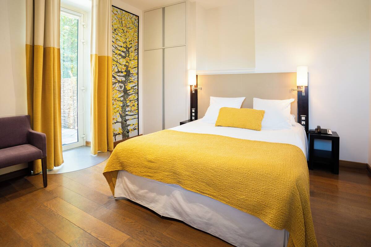 A hotel room with a large bed, a nightstand, a chair, and a view of the outdoors through a window with curtains.