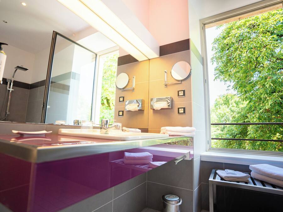 A modern bathroom with a large mirror, two sinks, and a window with a view of greenery.