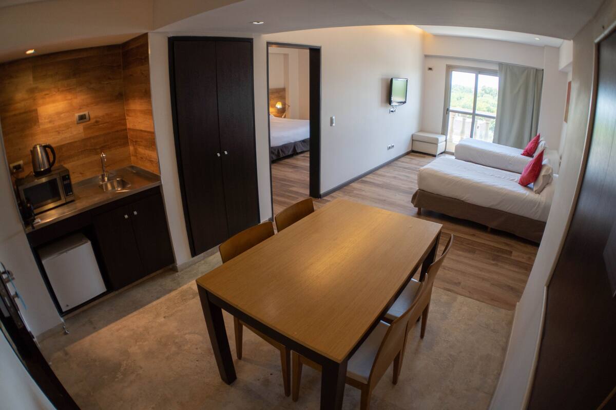 Ein kleines Hotelzimmer mit Küchenzeile, Essbereich, Bett und einem an der Wand befestigten Fernseher.