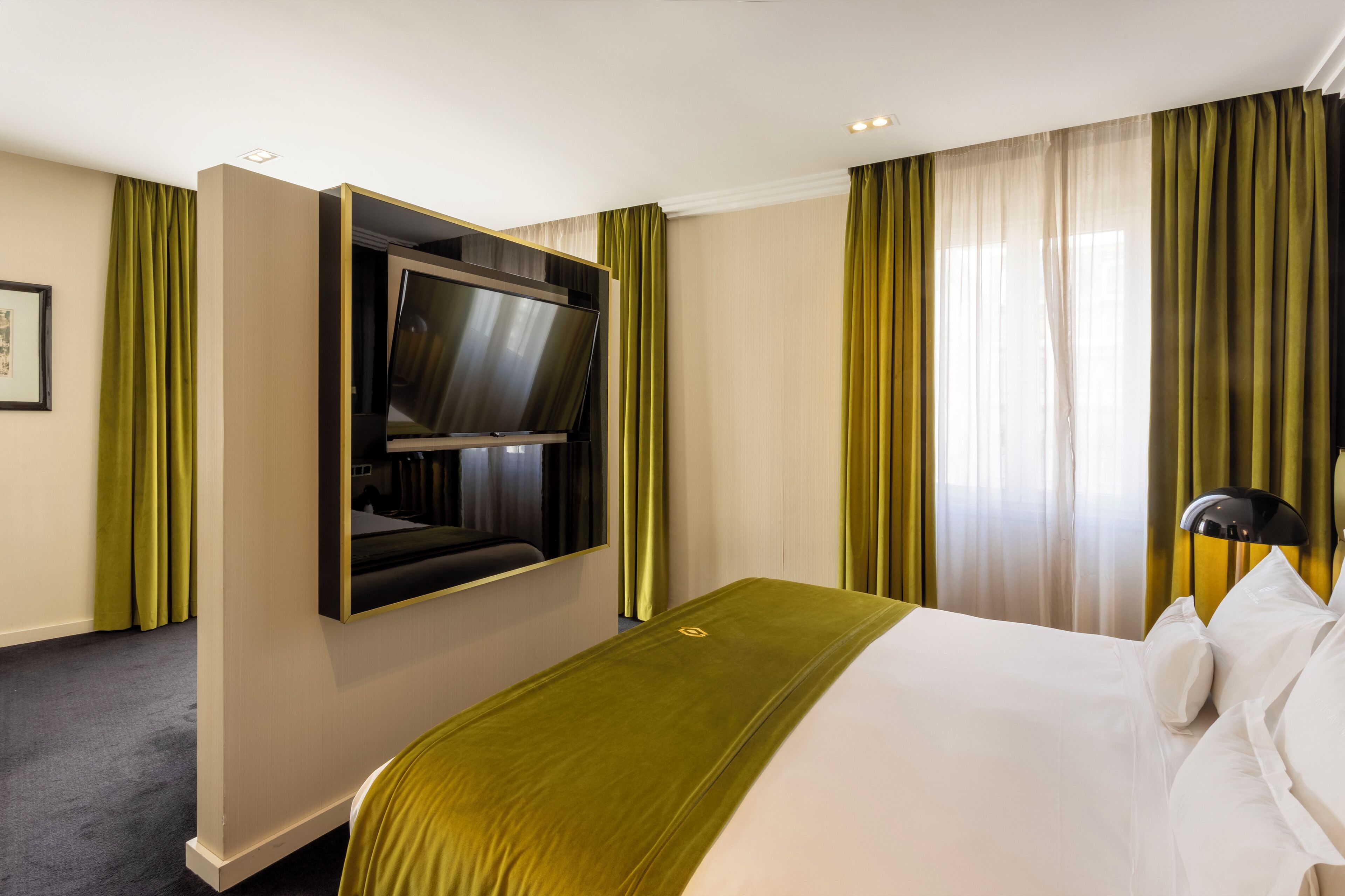 Una habitación de hotel con cama, televisión montada, lámpara y cortinas verdes.