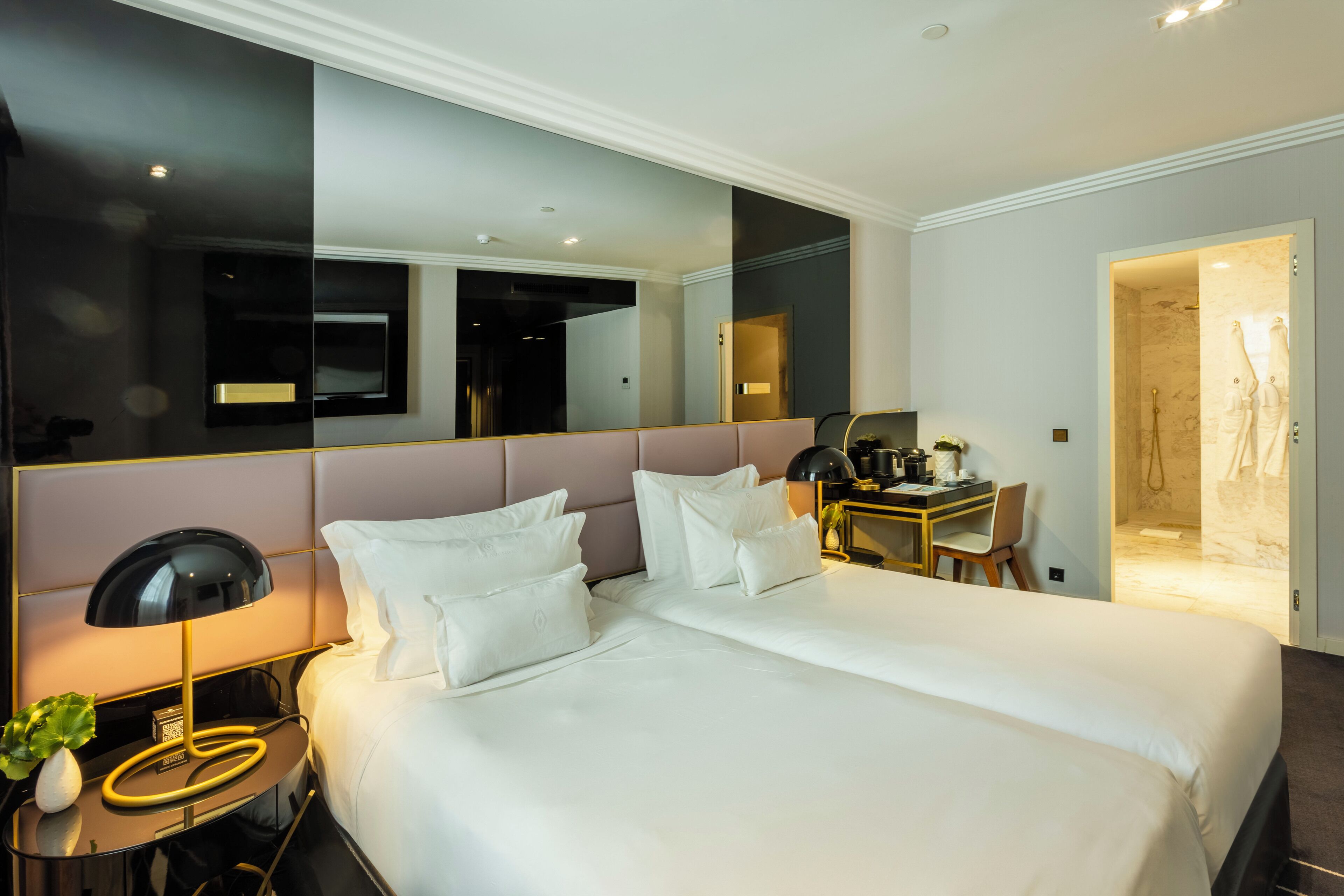 A modern hotel room with a large bed, bedside table, lamp, and a desk with a chair.