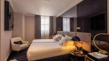 A hotel room with two beds, a chair, a desk with a lamp, a TV, and a large mirror.