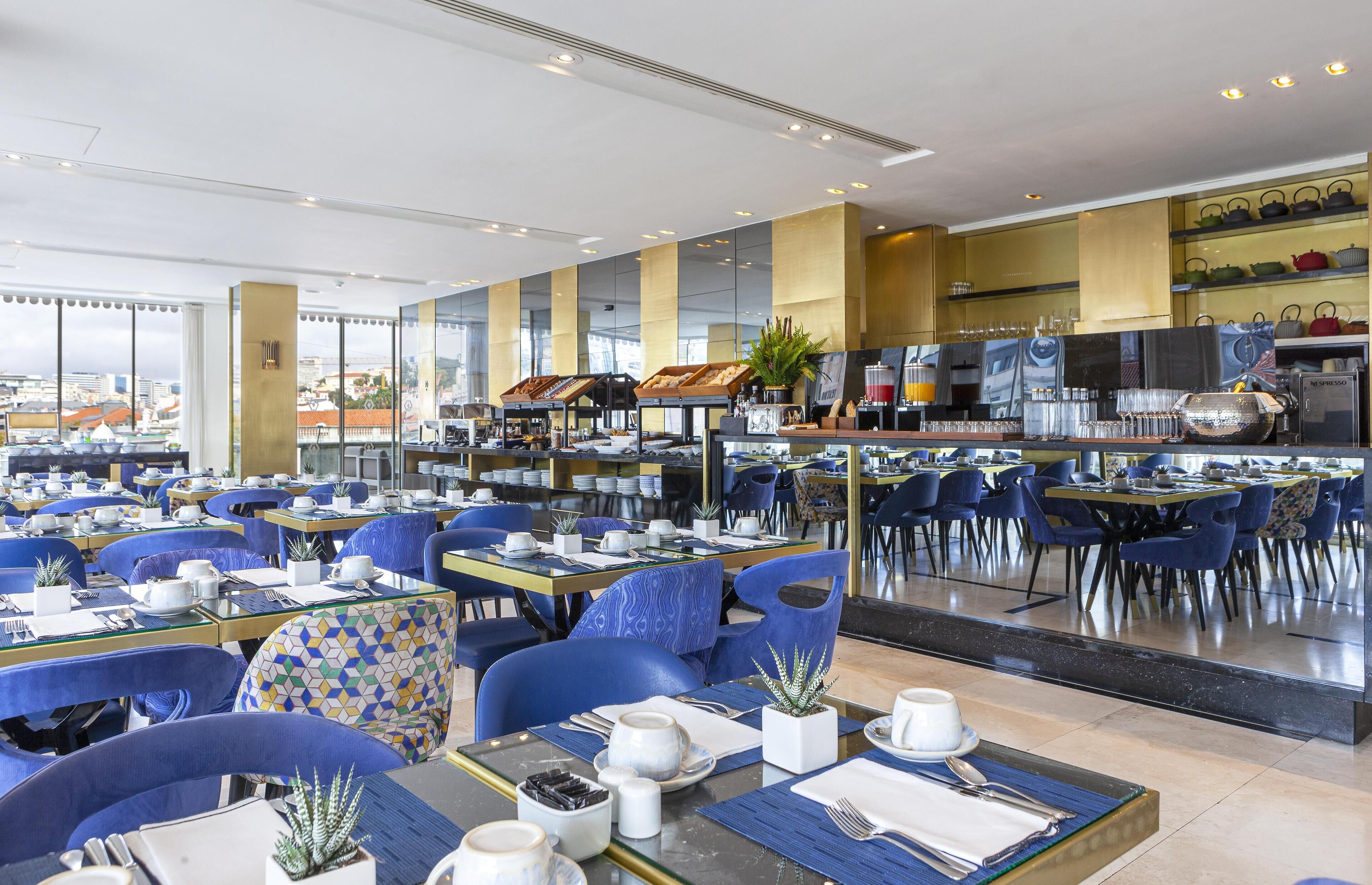A modern dining area with multiple tables and chairs, a buffet setup, and a view of the city through large windows.