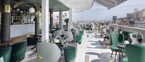 A rooftop bar with green upholstered chairs and tables, a bar area with a marble counter, and a city view.