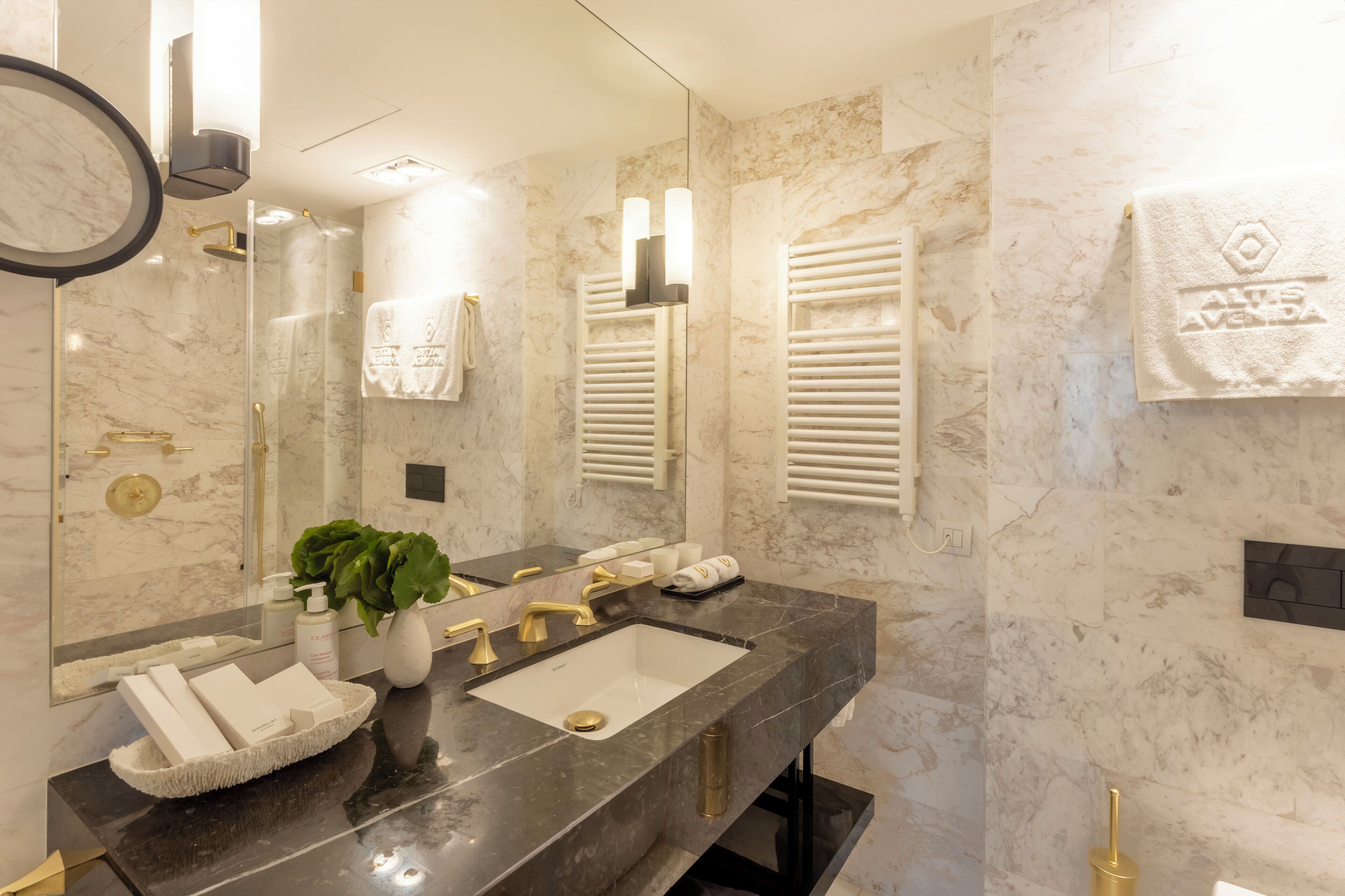 A modern bathroom with a marble sink, gold fixtures, a large mirror, and a towel rack with a branded towel.