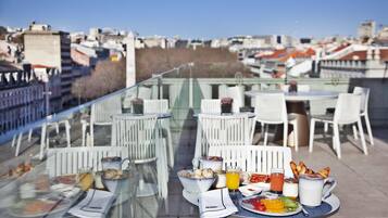Un área de comedor en la azotea con mesas y sillas, una mesa de vidrio y vistas a la ciudad.